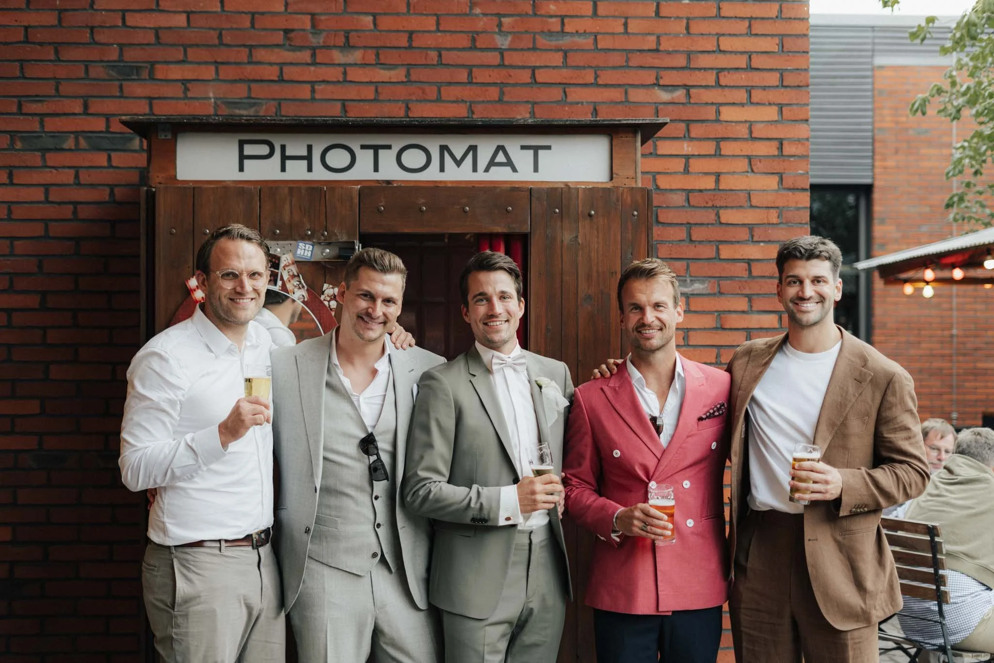 Eine Gruppe von sechs Männern steht vor einem Fotokabinengebäude, lächelt und hält Getränke in der Hand, bei einer Veranstaltung im Freien.