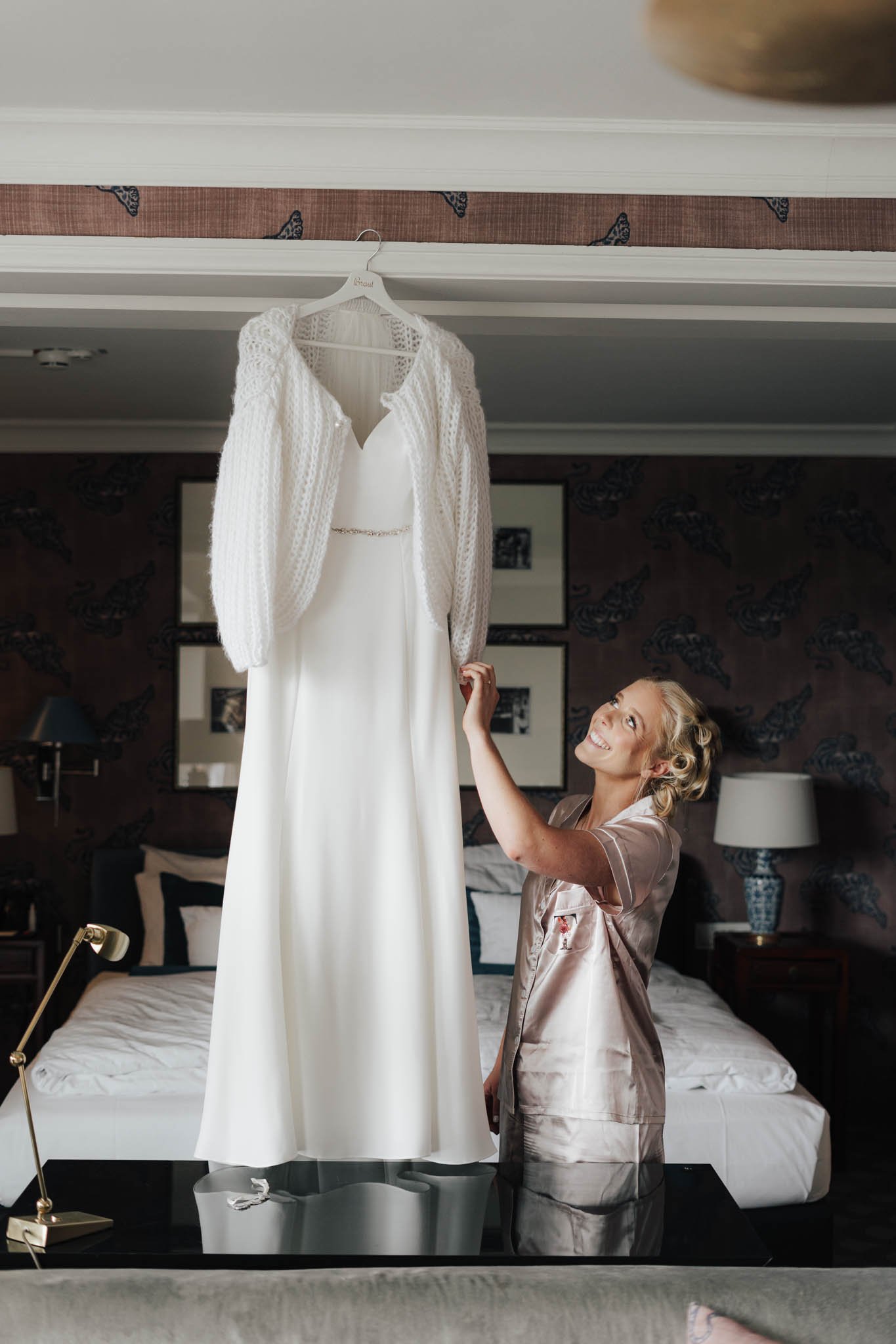 Eine braut in einem Schlafzimmer betrachtet ein Hochzeitskleid, das an einer Deckenstange hängt. Sie trägt ein Satin-Nachtwäsche-Set mit einem Lächeln auf dem Gesicht.