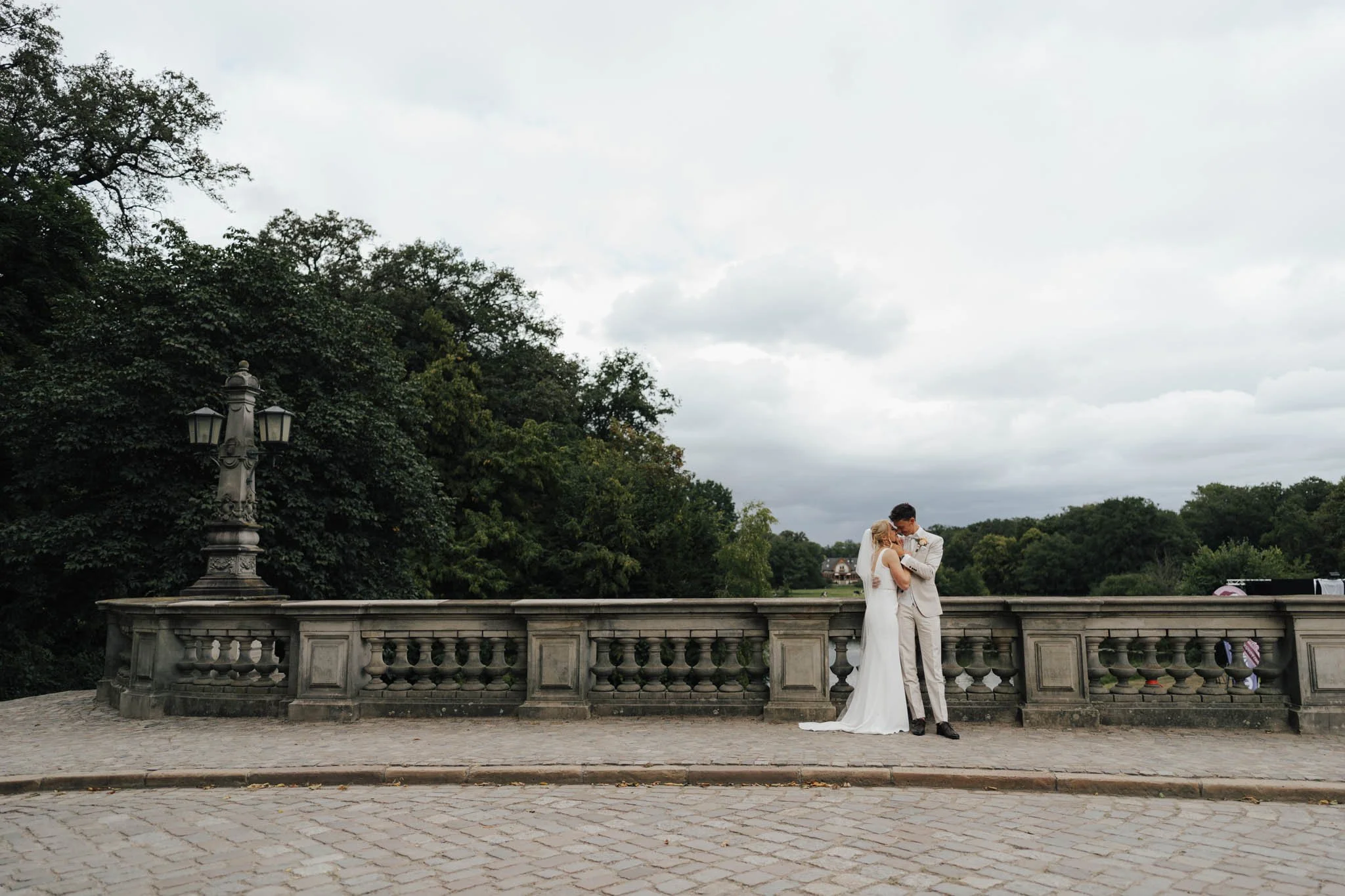 Ein Brautpaar umarmt sich auf einer Brücke im Bürgerpark Bremen, umgeben von Bäumen, unter einem bewölkten Himmel.