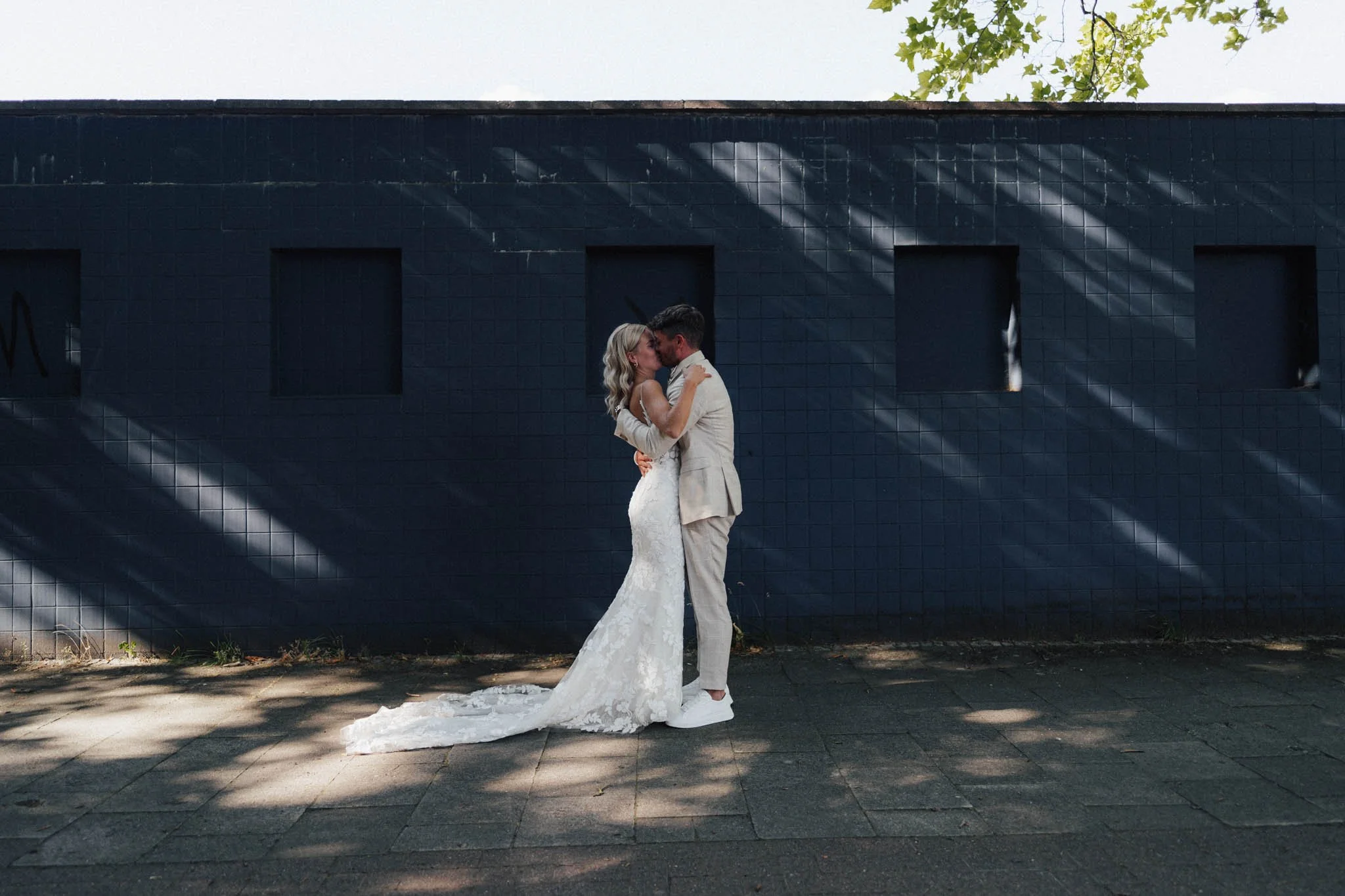 Ein frisch verliebtes Brautpaar umarmt sich und küsst sich vor einer schwarzen Wand mit vier unbedeckten Fenstern, während Sonnenstrahlen Schatten auf die Wand werfen.