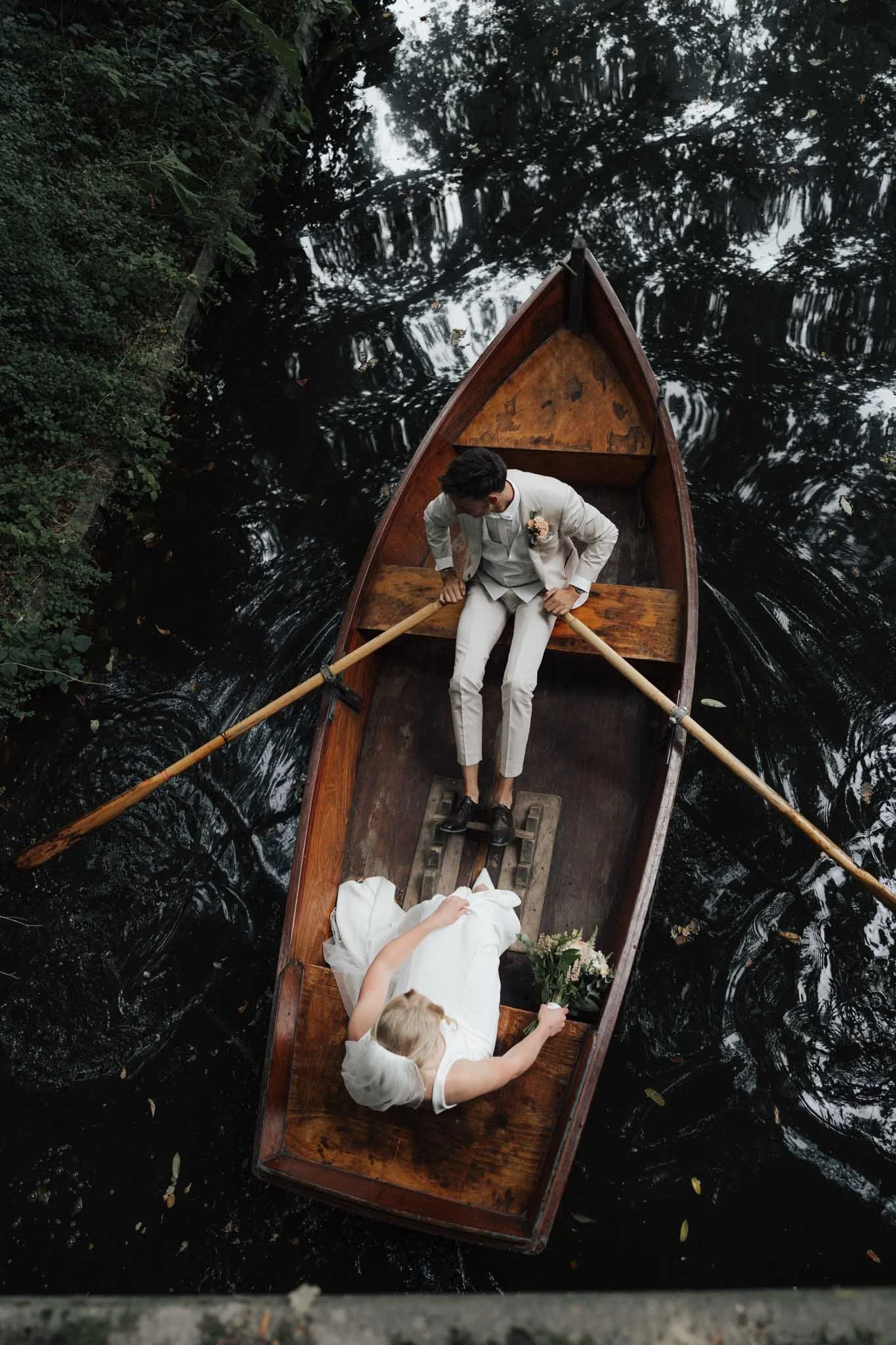 Eine Braut sitzt in einem Holzboot auf einem Fluss, während der Bräutigam am Ruder sitzt. Die Fotografie wurde von oben aufgenommen.