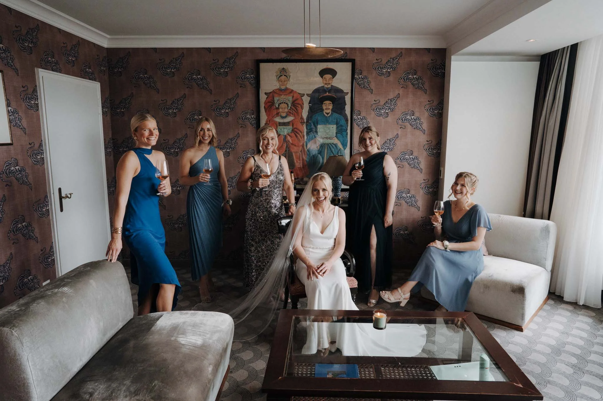 Sieben Frauen in eleganter Abendkleidung bei einer Feier im Wohnzimmer, wir sechs halten Wein, eine sitzt auf einem Stuhl, alle lachen, im Hintergrund eine Wand mit Tapete, ein großes Gemälde und ein Couchtisch mit Kerze.