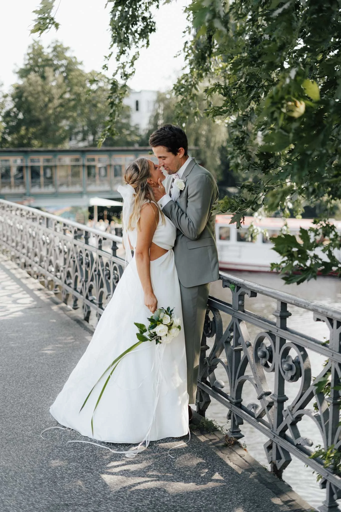 Ein Brautpaar auf einer Brücke am Fluss in Hamburg, umarmend bei schönstem Licht, mit Bäumen im Hintergrund.