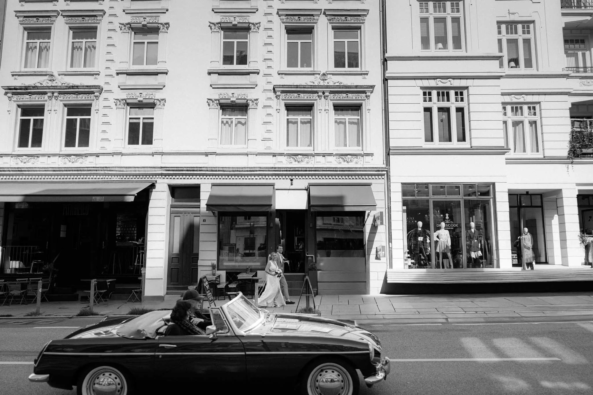 Schwarzes Cabriolet fährt an einem Stadtstraßenrand vorbei, im Hintergrund mehrstöckige Altbauten in Hamburg mit Geschäften und Schaufenstern, eine Frau in Brautkleid läuft auf dem Bürgersteig.
