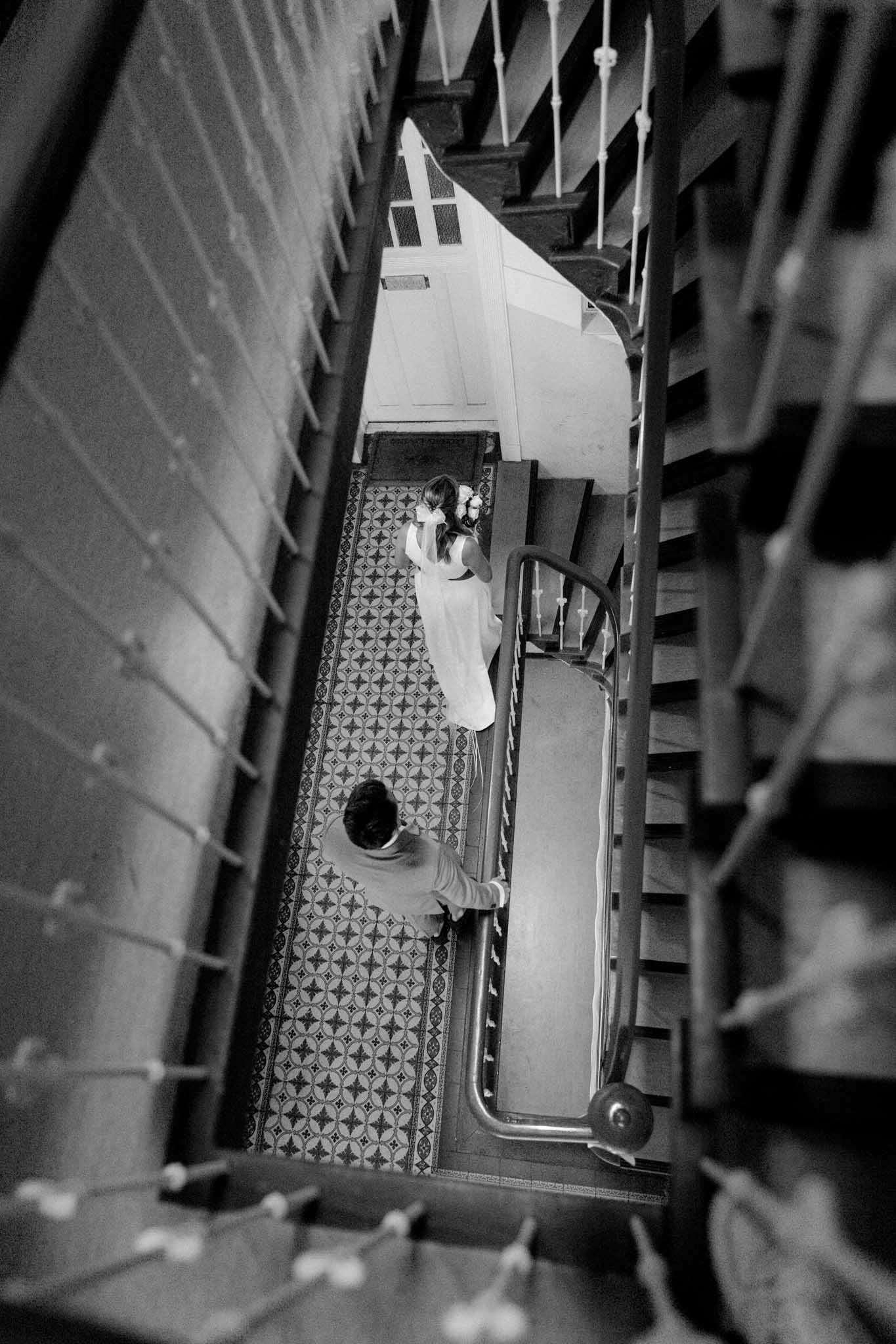 Blick von oben in eine Treppenhaus, mit einer jungen Frau in einem weißen Kleid und einem Mann, die sich gegenüberstehen.