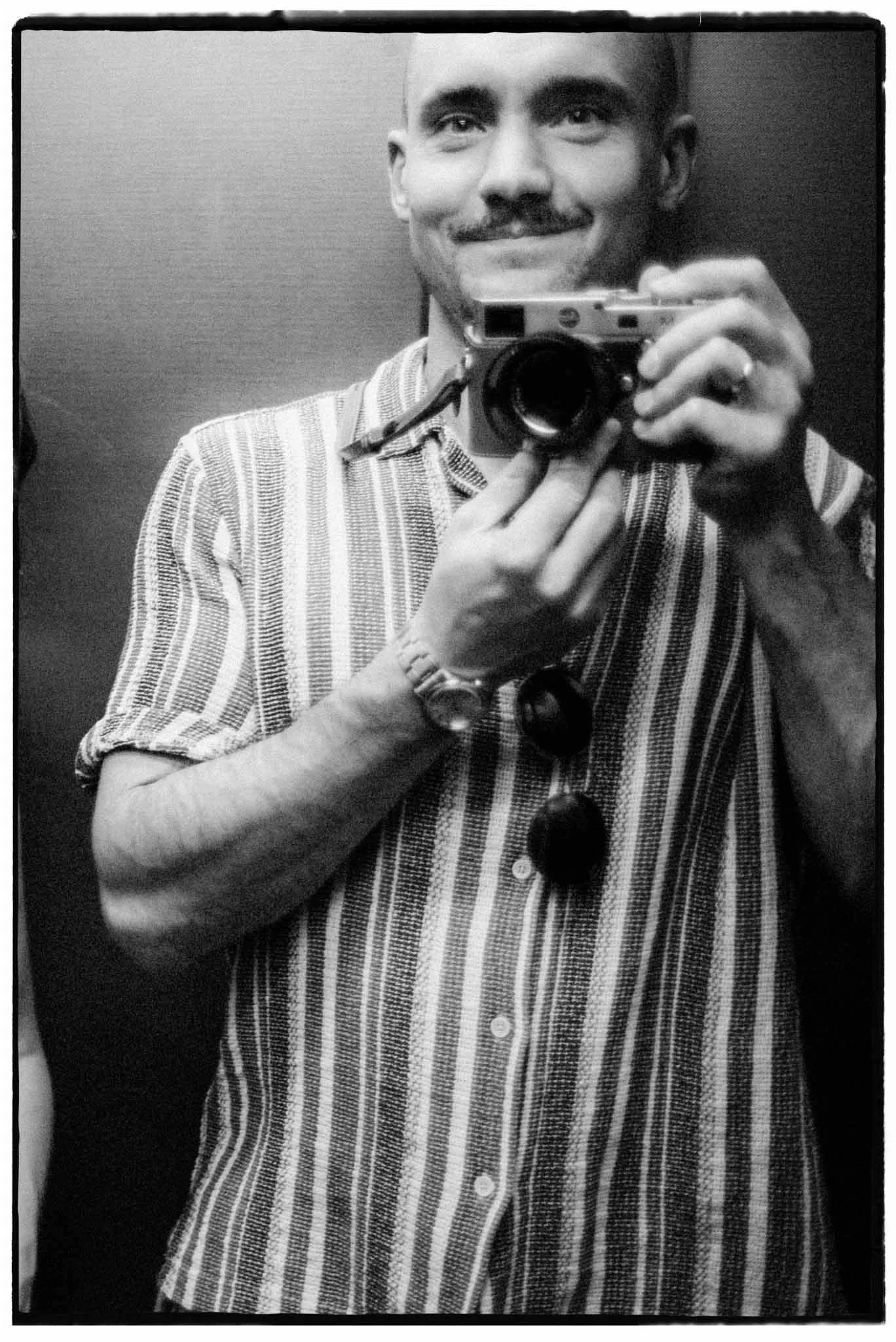 Spiegelbildfotofgrafie in schwarz und weiß eines Mannes in gestreiftem Hemd mit Kamera in der Hand und Sonnenbrille im Hemd.