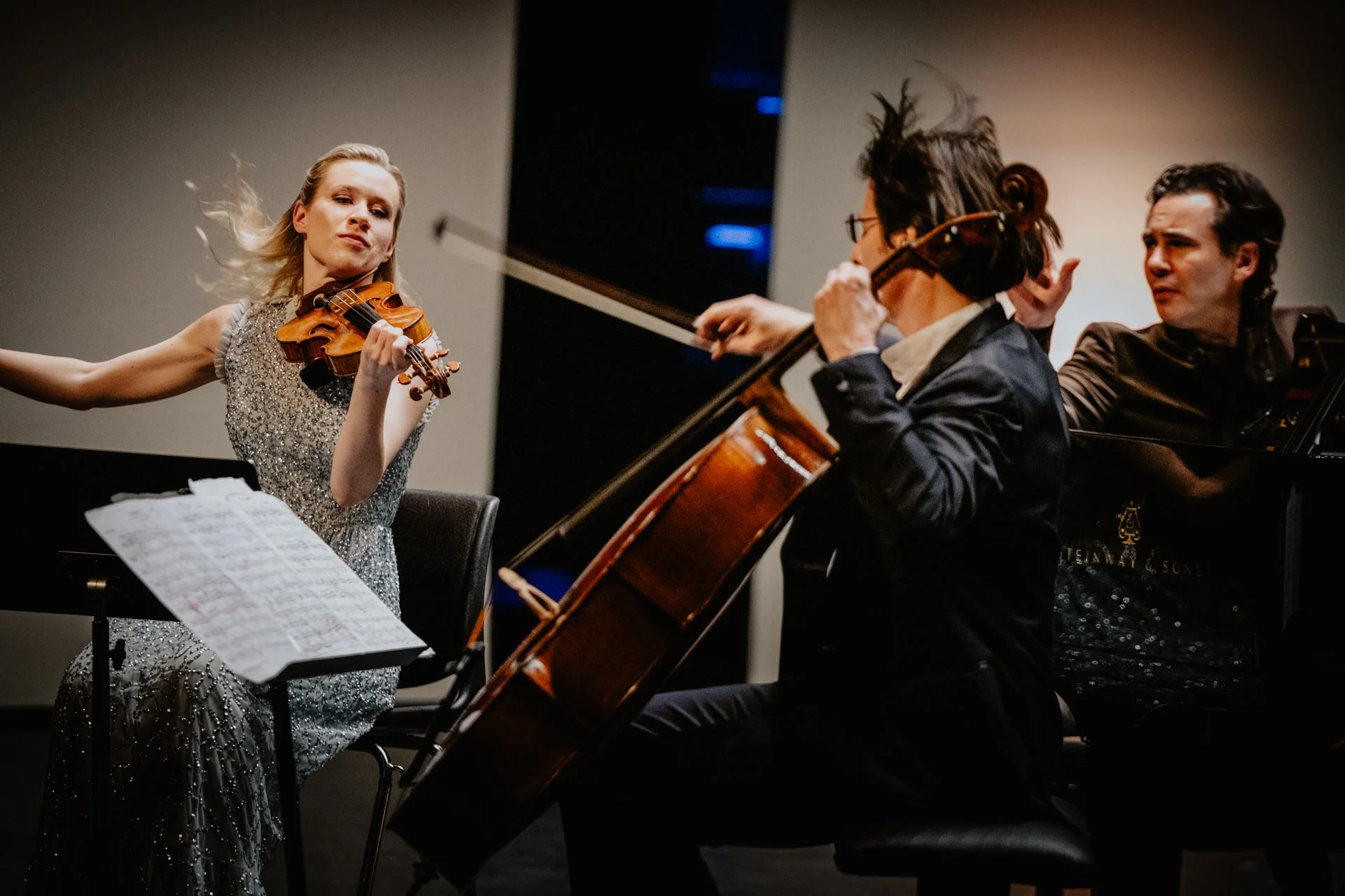 Three musicians perform together, a woman playing the violin, a man playing the cello, and another man playing the piano.