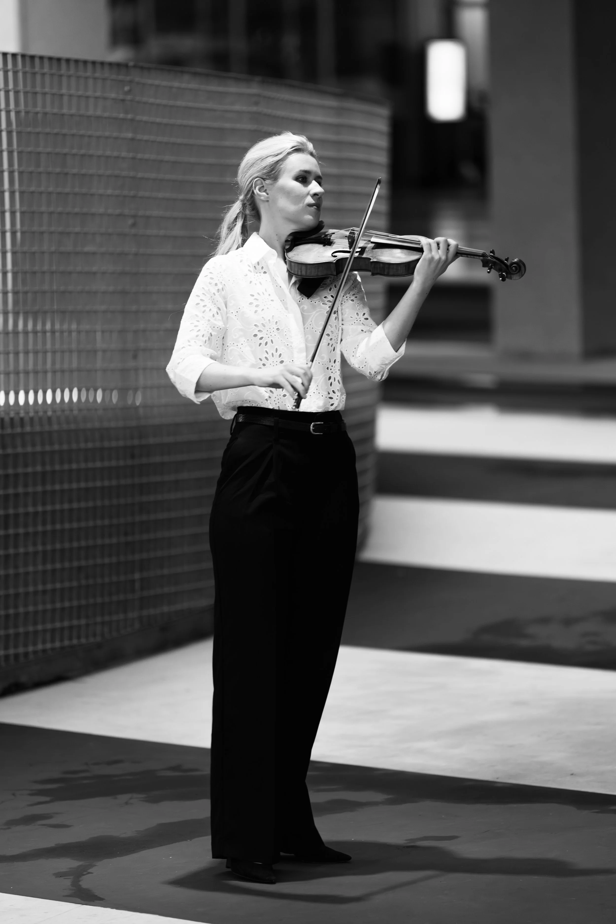 Black and white photo of a Eldbjørg Hemsing playing the violin, standing outdoors with a modern building in the background.