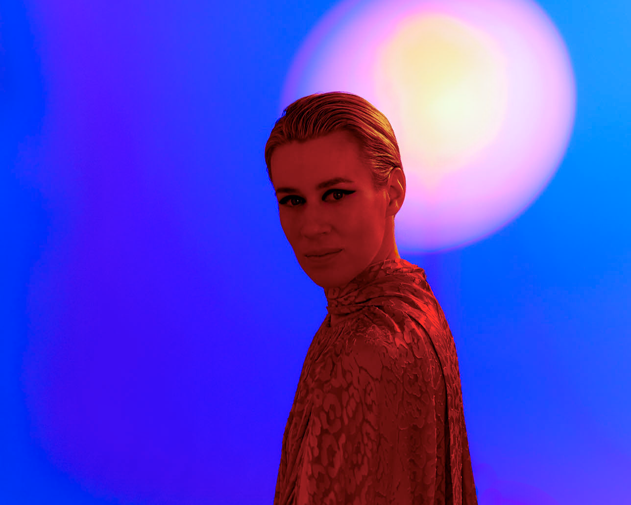 A woman with slicked-back blonde hair and wearing a red patterned top stands in front of a vibrant background with blue and purple lighting and a large glowing orb.