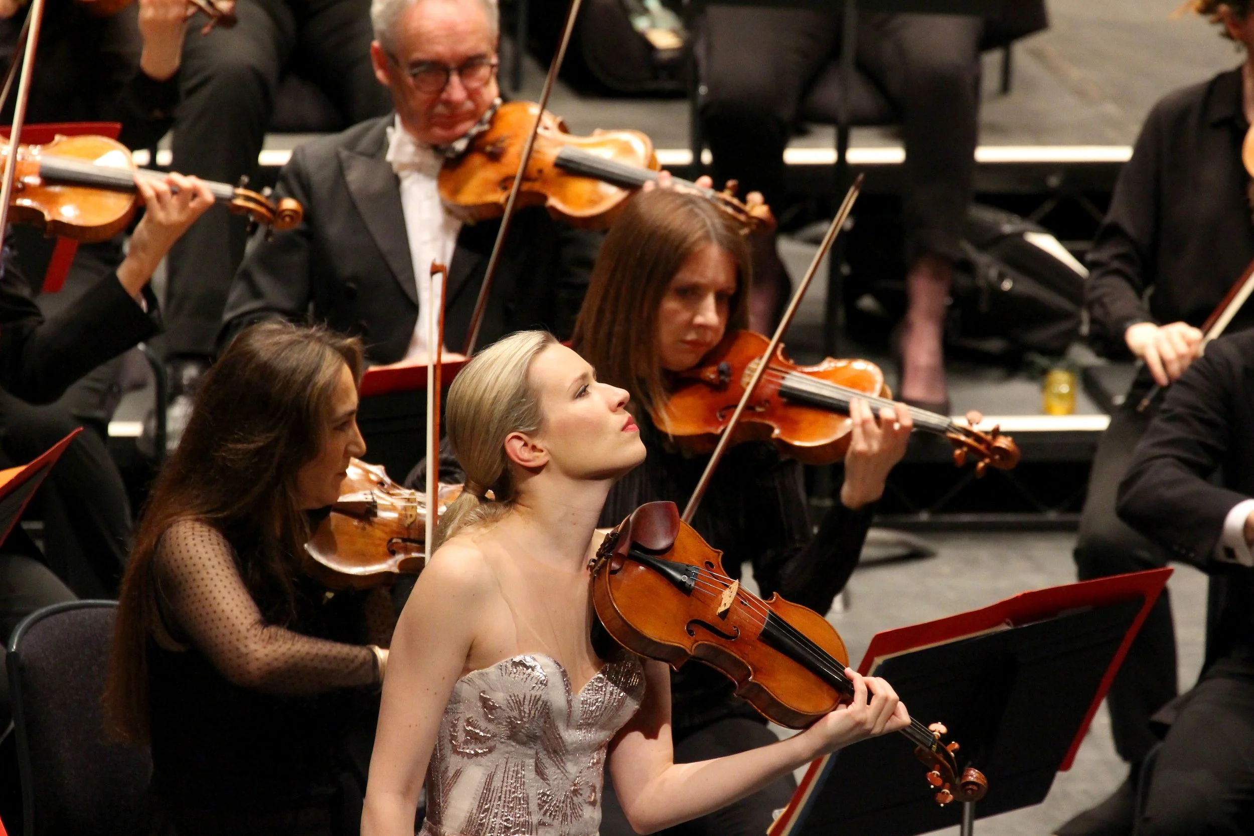 A group of musicians playing violins in an orchestra performance, with a woman in a strapless dress in focus.