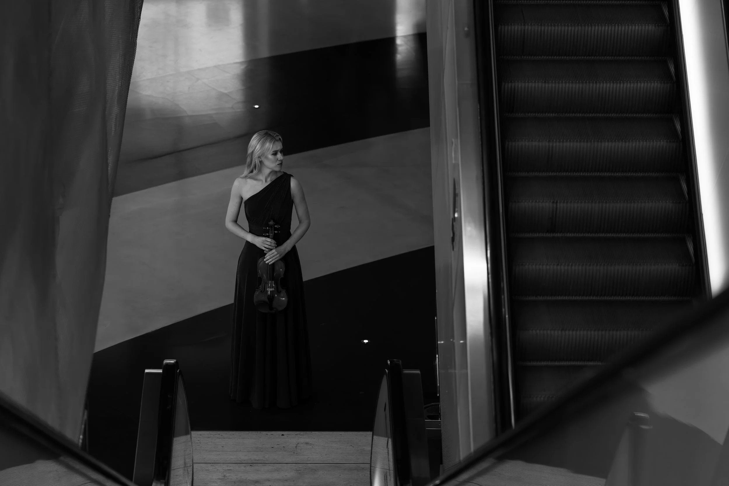 A woman in a long, elegant black dress holding a violin, standing alone near an escalator in a modern, minimalistic interior space.