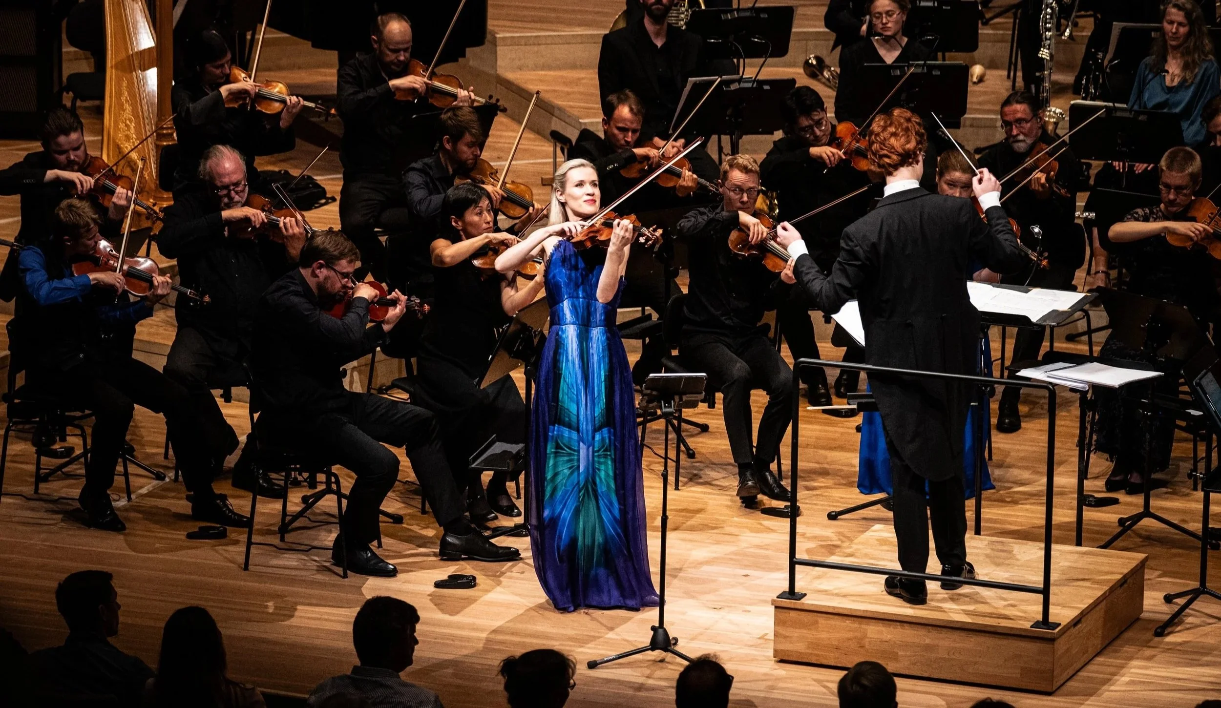 Orchestra performing on stage with a female solo violinist in a blue dress and a conductor in black tuxedo leading.