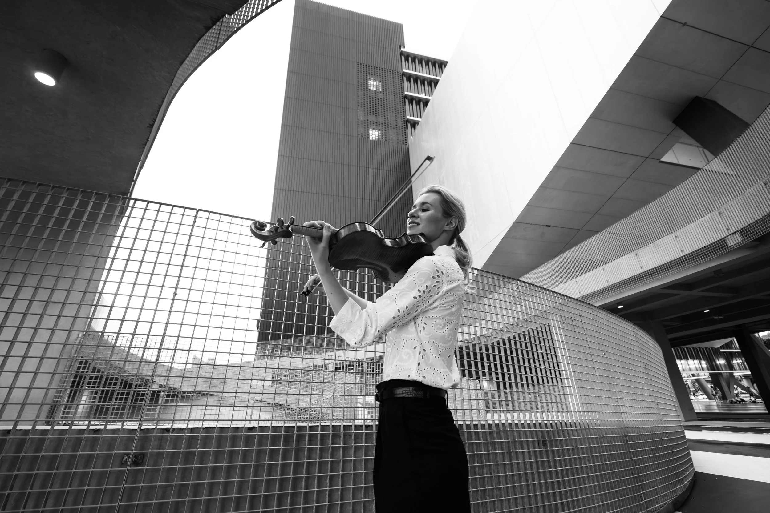 A woman playing violin outdoors in an urban setting with modern buildings and a curved metal fence in the background.