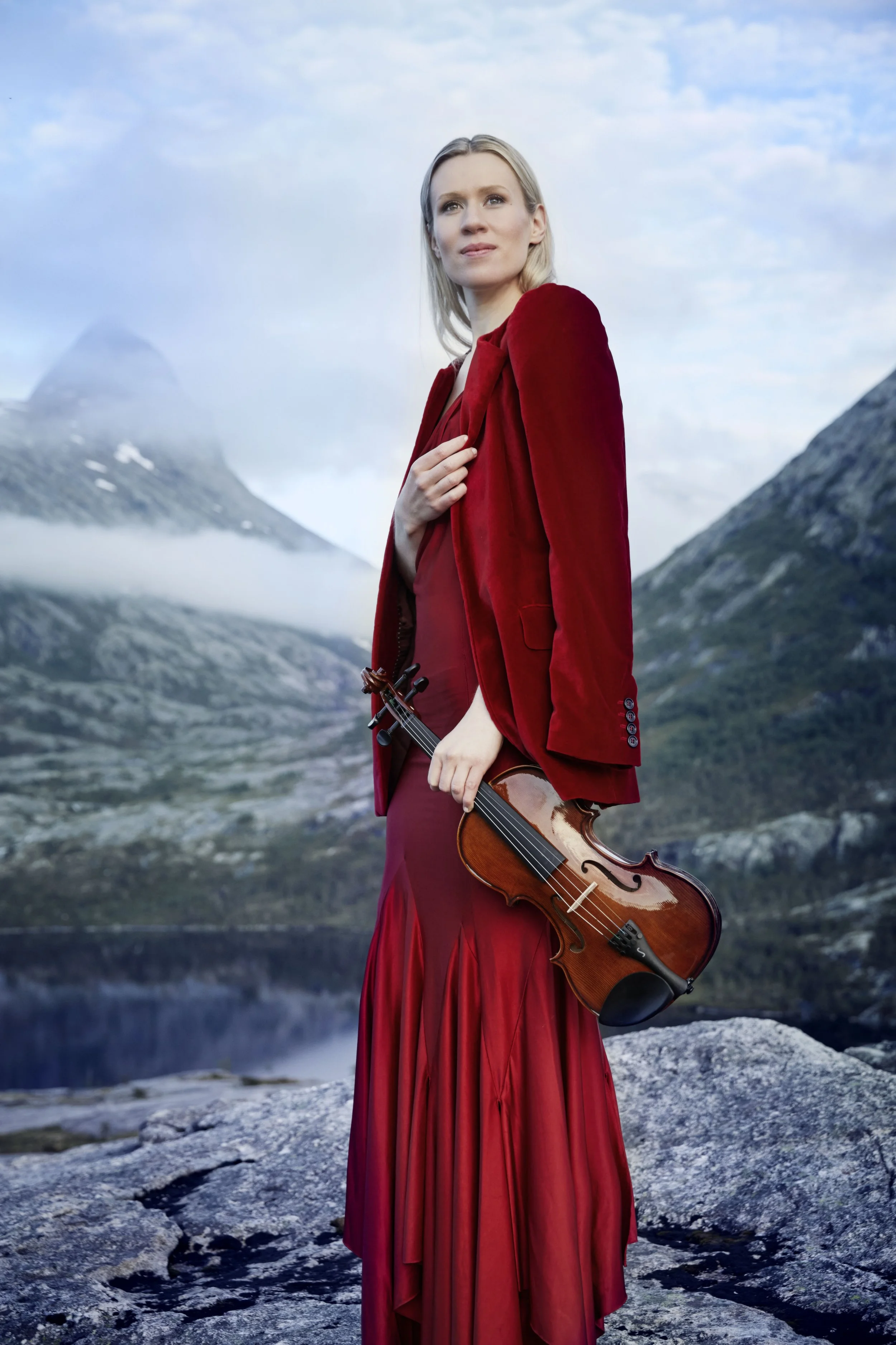 Eldbjørg Hemsing in a long red dress and matching red blazer stands outdoors on rocky terrain near a lake with mountains and cloudy sky in the background, holding a violin.