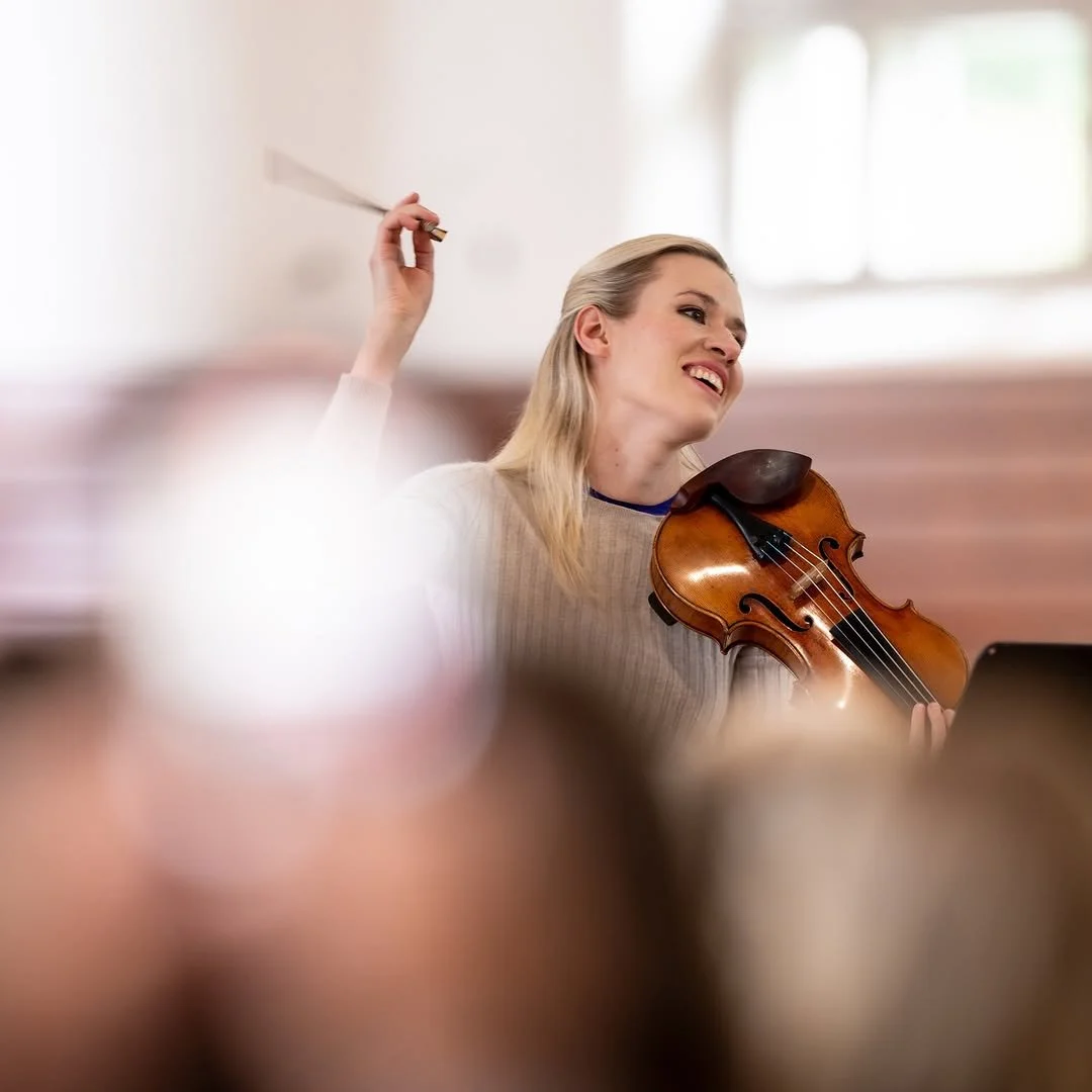 A woman playing a violin during a music rehearsal or performance, with a smile on her face and a music stand in front of her.