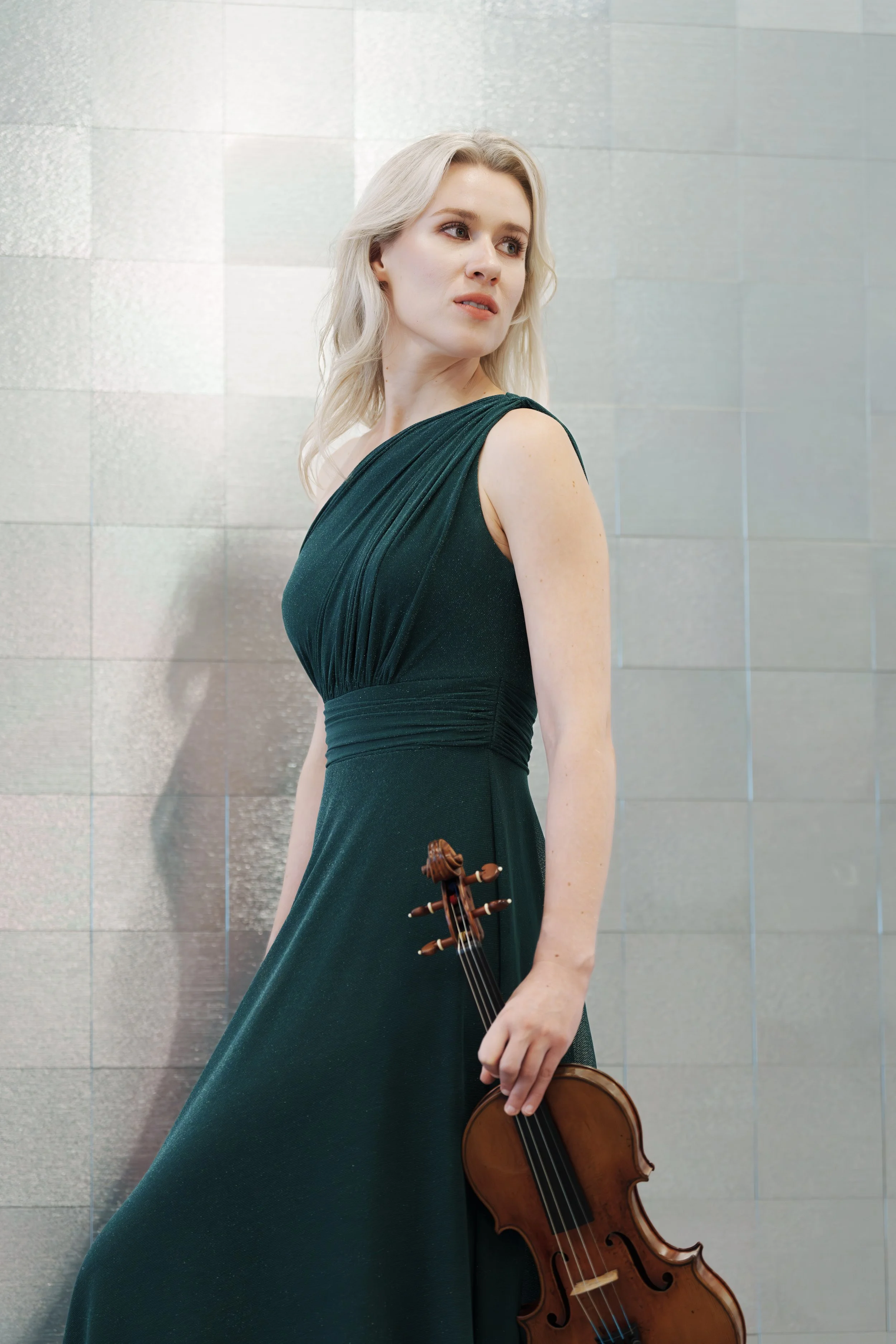 A blonde woman in a dark green evening gown holding a violin, standing against a metallic tiled wall.