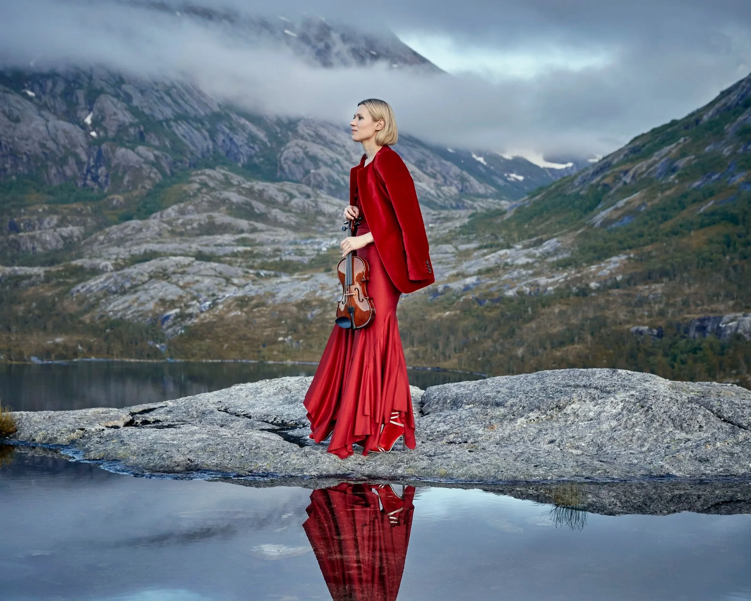 A woman in a long red dress and matching red blazer holding a violin walks on a rock beside a reflective body of water, with mountains and cloudy skies in the background.