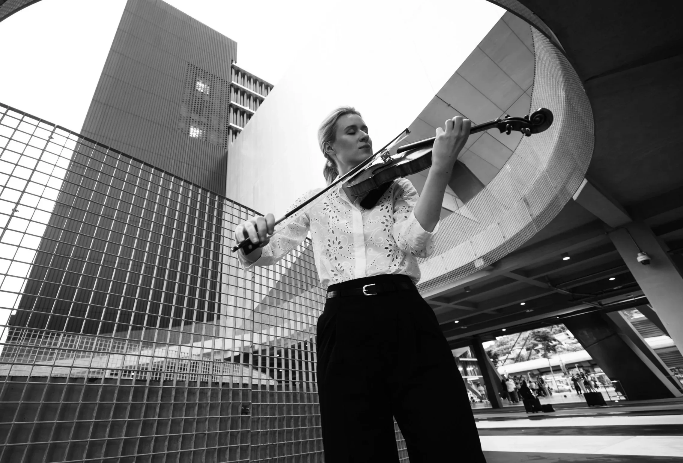 A woman playing the violin outdoors in an urban setting with modern architecture around her.