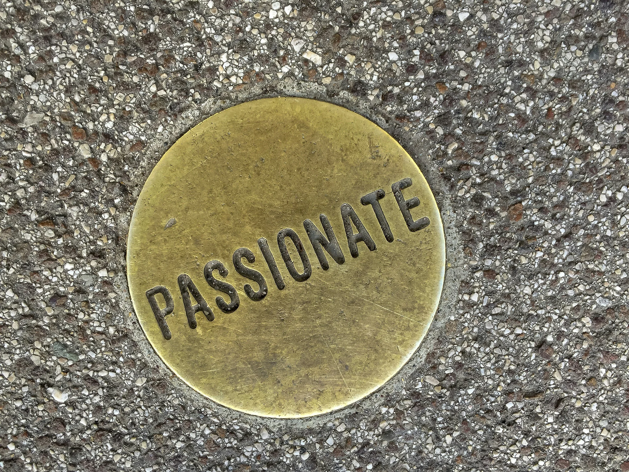 Close-up of a round brass plate embedded in concrete pavement, with the word 'PASSIONATE' engraved in black.