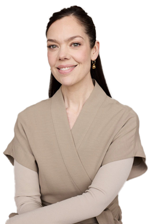 Une femme souriante avec des cheveux bruns attachés, portant une tenue beige et un haut gris clair, posant devant un fond noir.