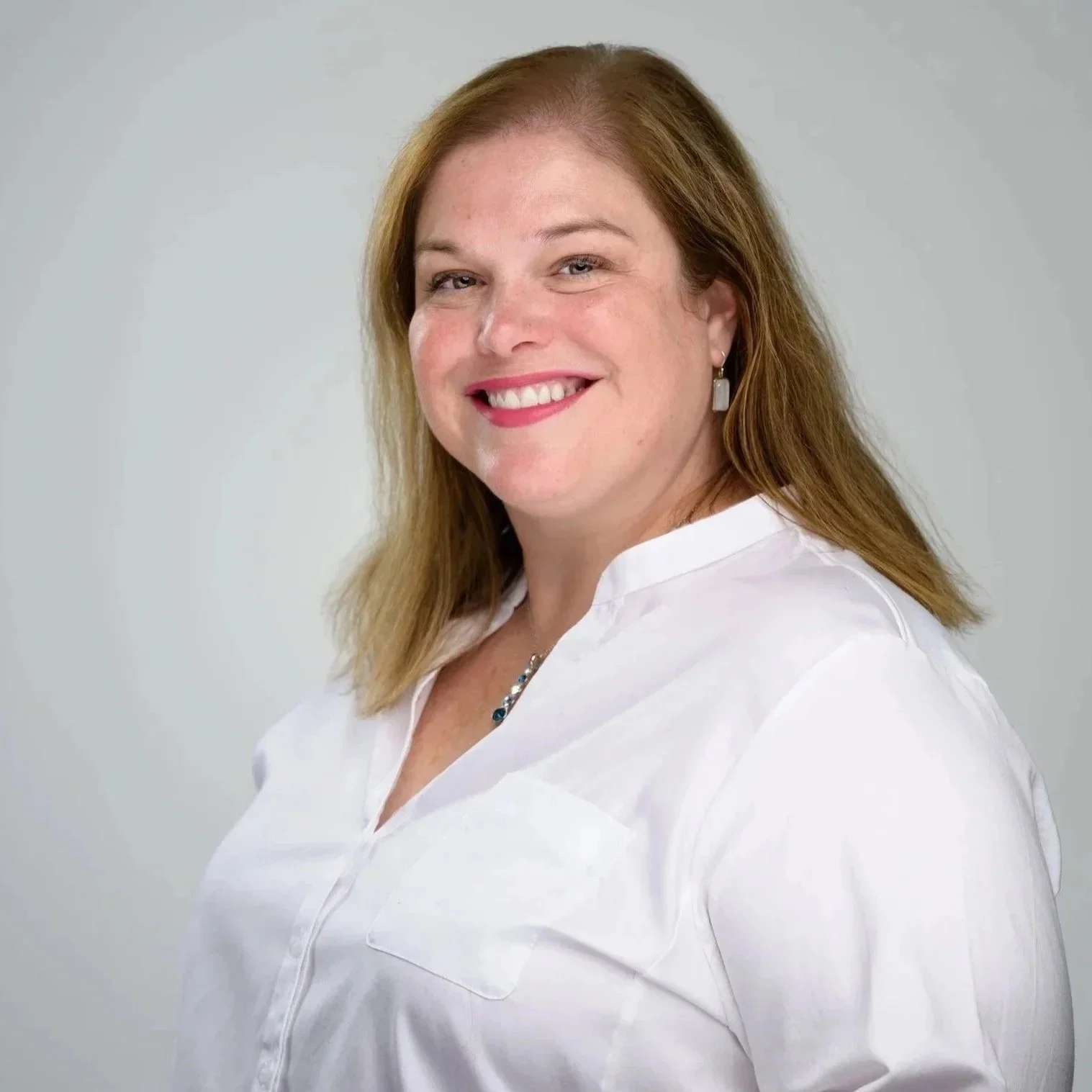 A smiling woman with shoulder-length light brown hair, wearing a white button-up shirt and earrings, posing against a plain light gray background.