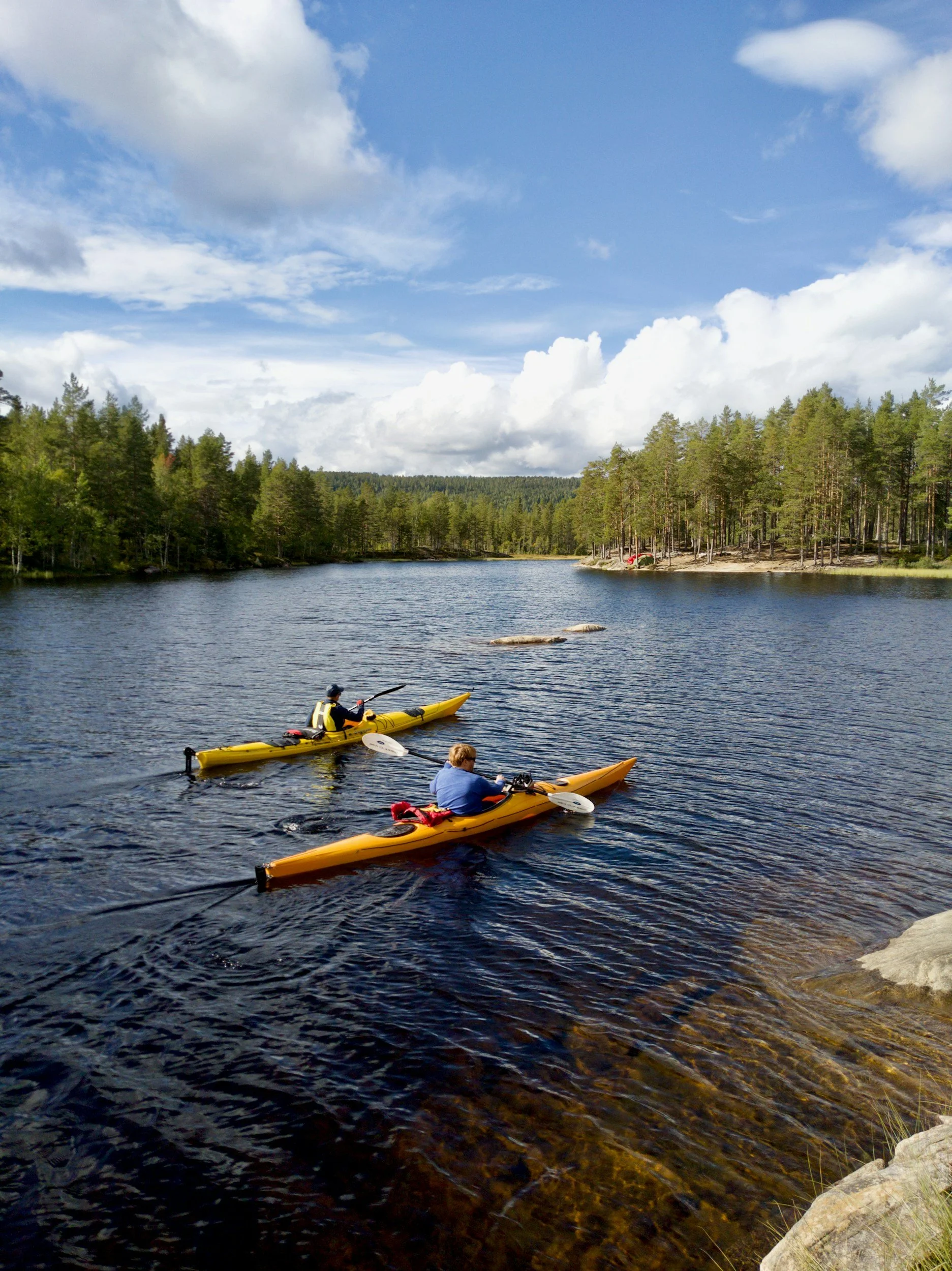 Two people kayaking on a lake with a forested shoreline and cloudy sky in the background.