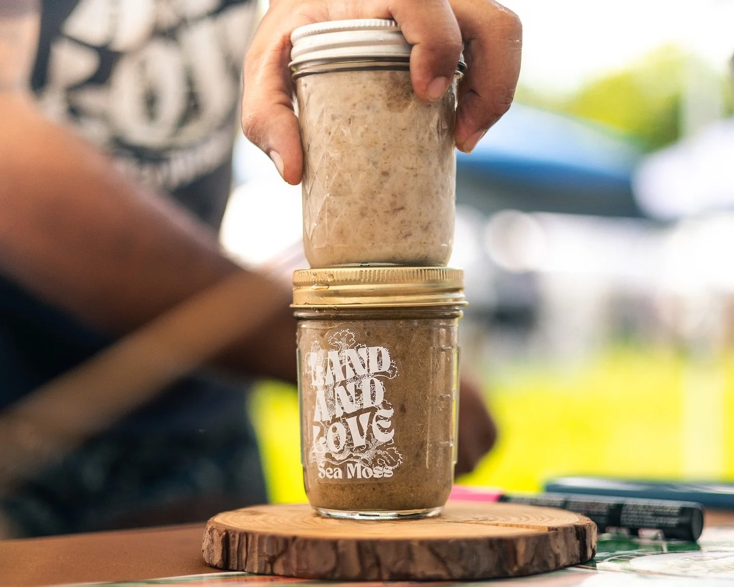 A hand holding a jar labeled 'Sea Moss' stacked on top of another jar with a light colored substance inside, both jars placed on a wooden surface.