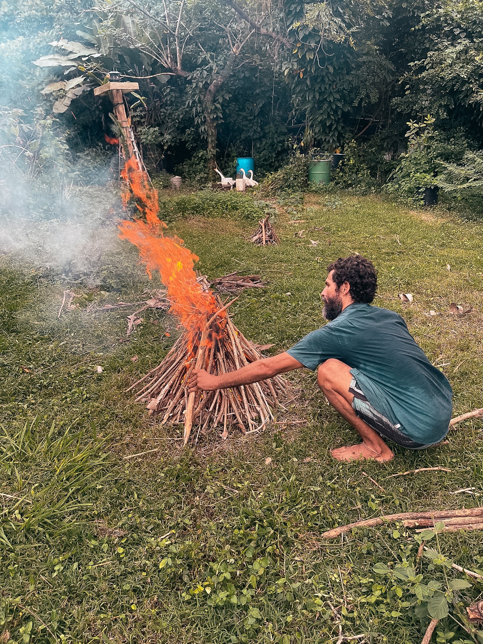 A man crouching beside a small bonfire made of sticks, tending to the flames in a backyard with grass, trees, and garden items in the background.