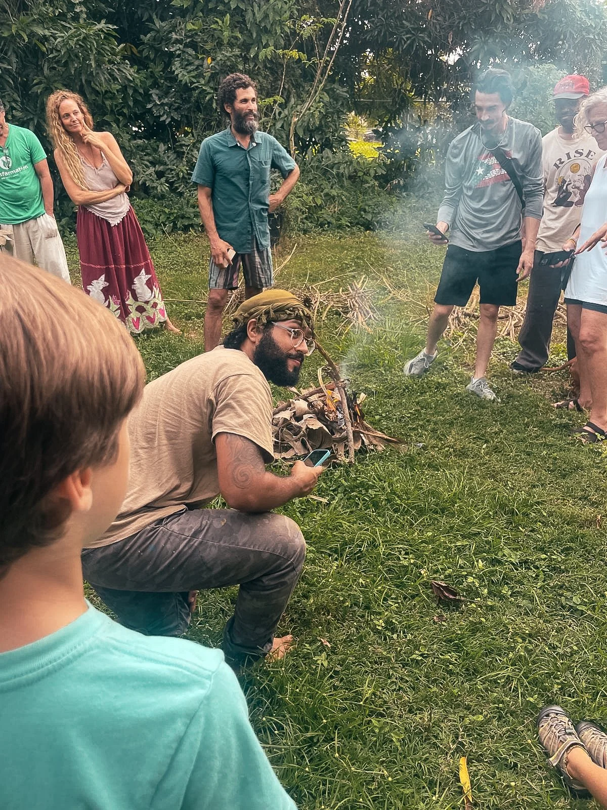 Group of people gathered outdoors near a small campfire in a green, wooded area, some standing and others sitting, engaged in conversation.