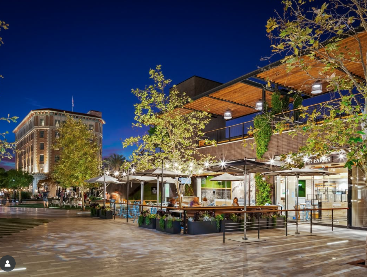 Outdoor dining area at dusk with tables, umbrellas, and people, in a modern building with a wooden roof extension and greenery, in an urban setting.