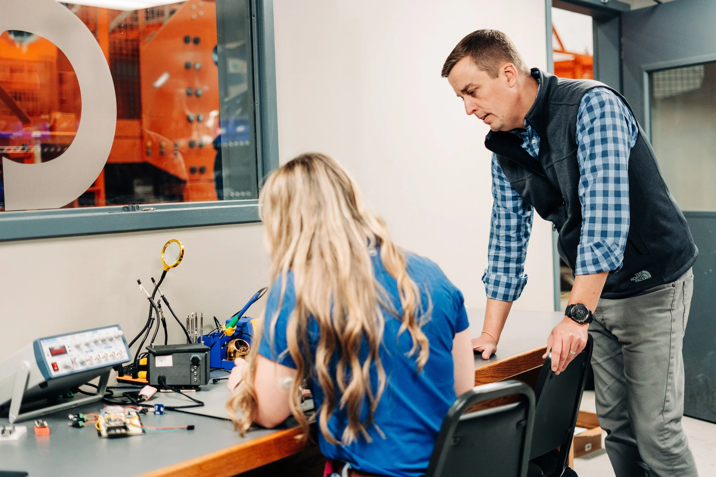 A man standing over a girl seated at a workbench with electronics and tools, in a workshop or classroom.