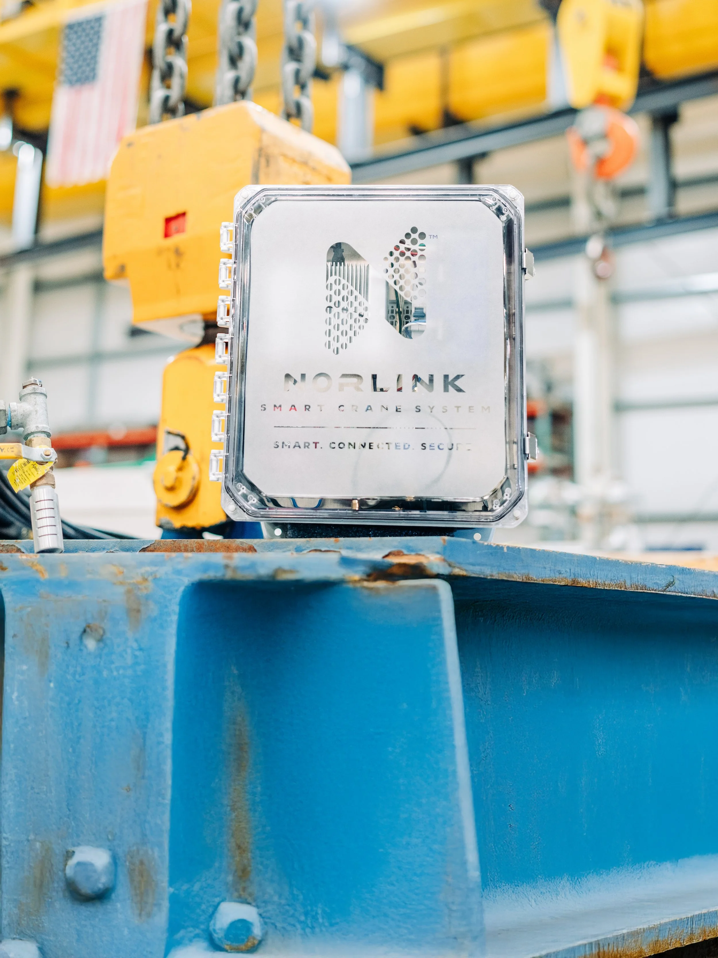Close-up of a Norlink Smart Crane System control box mounted on aged blue steel, with yellow industrial equipment and hooks in the background.