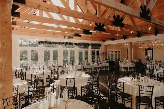 Event hall with wooden beams ceiling, white tablecloth-covered round tables, black chairs, and large windows, decorated for a celebration or wedding reception.