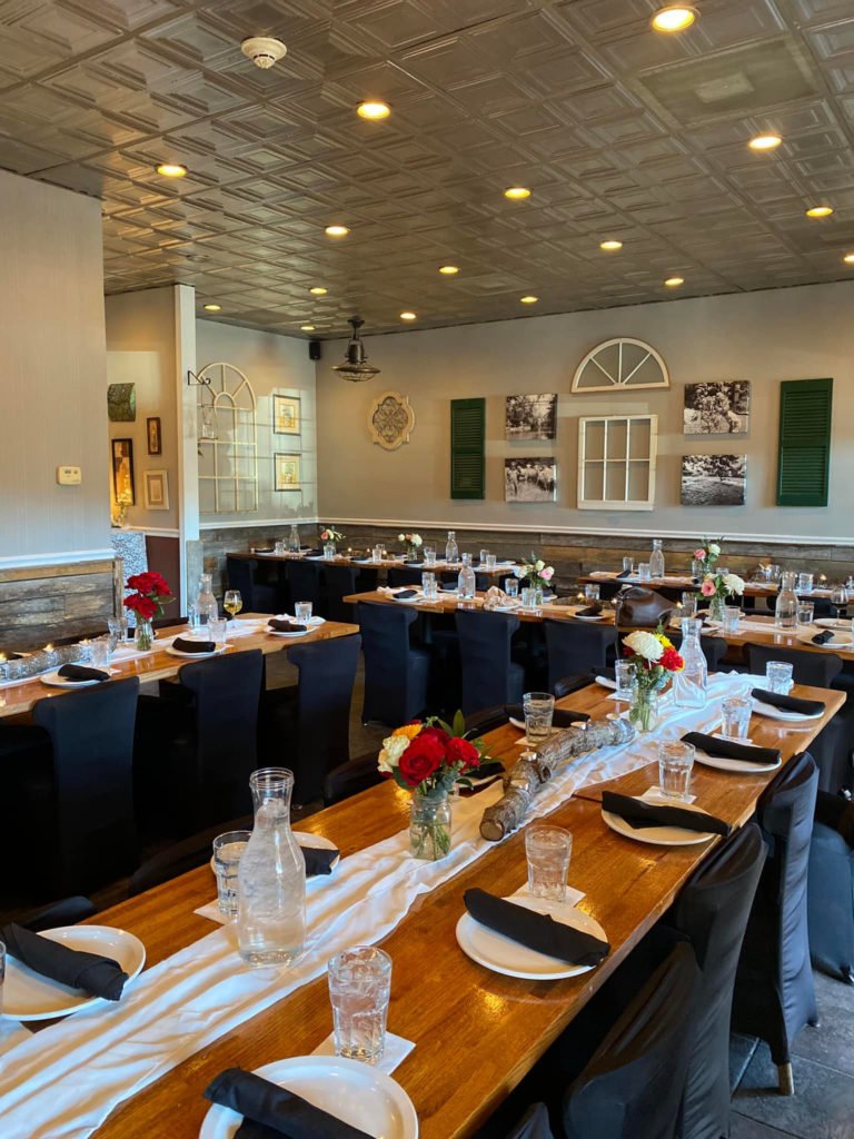 A decorated banquet room with long wooden tables set for a meal, featuring black napkins, white plates, water glasses, and small floral centerpieces in glass jars with red, white, and pink flowers. The room has a gray wall with decorative art and win