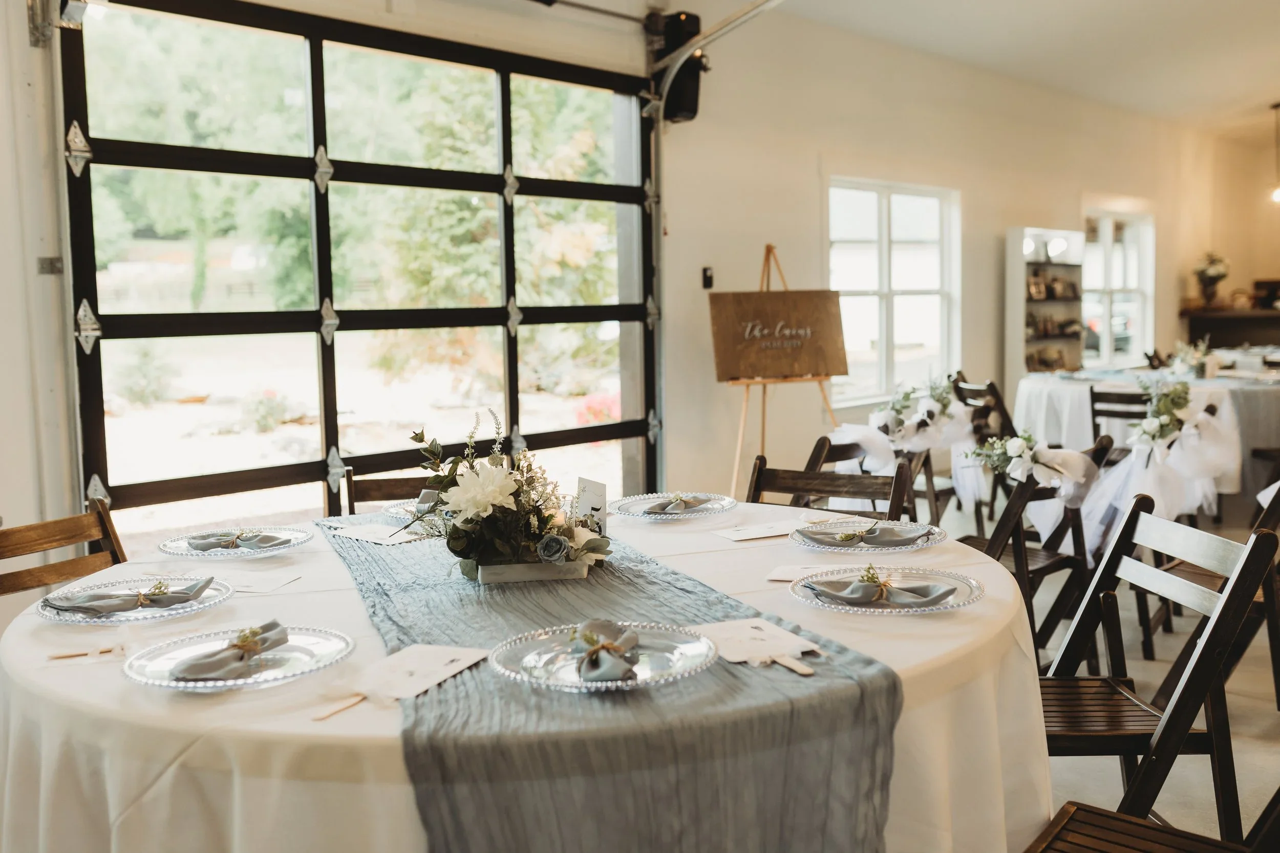A decorated banquet table set for a formal event, with a centerpiece of white flowers and candles, surrounded by white plates, napkins, and small decorative items, in a bright room with large windows and a garage door.