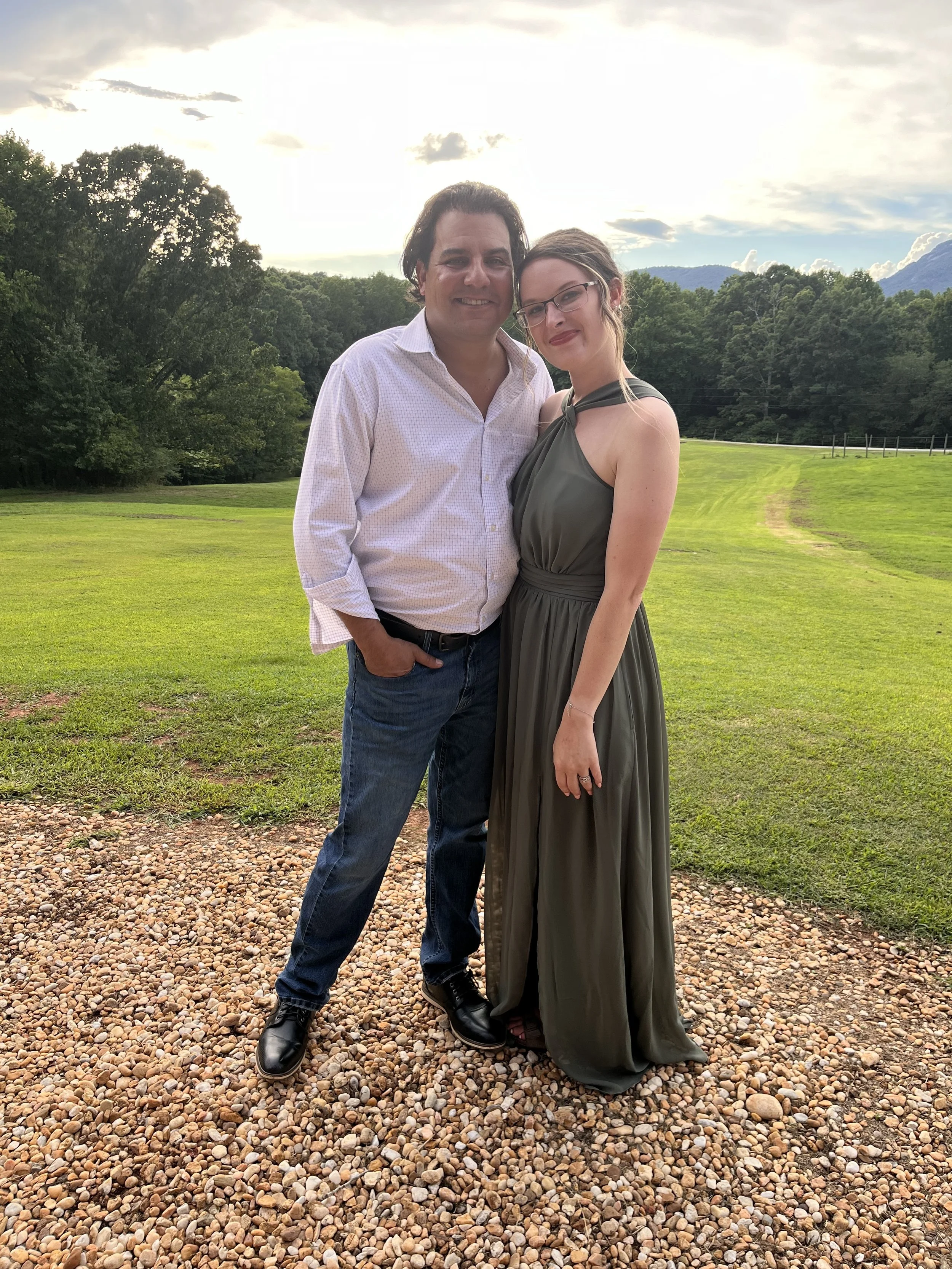 A man and woman standing outdoors on a gravel surface, with green grass and trees in the background, during sunset or evening.