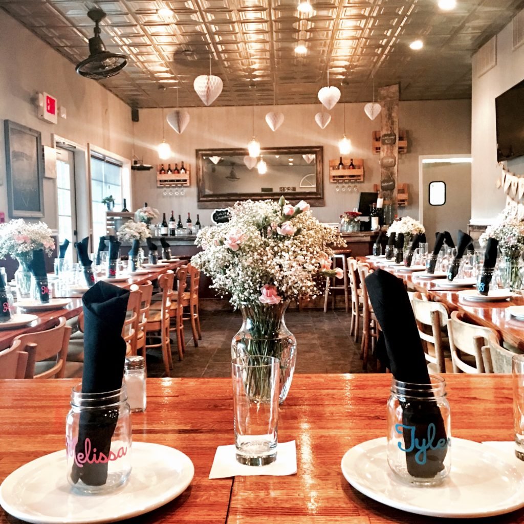 A decorated restaurant interior with a long dining table set with black napkins in mason jars with personalized names, and a prominent floral centerpiece of baby's breath and pink roses. Heart-shaped paper lanterns hang from the ceiling, and there ar