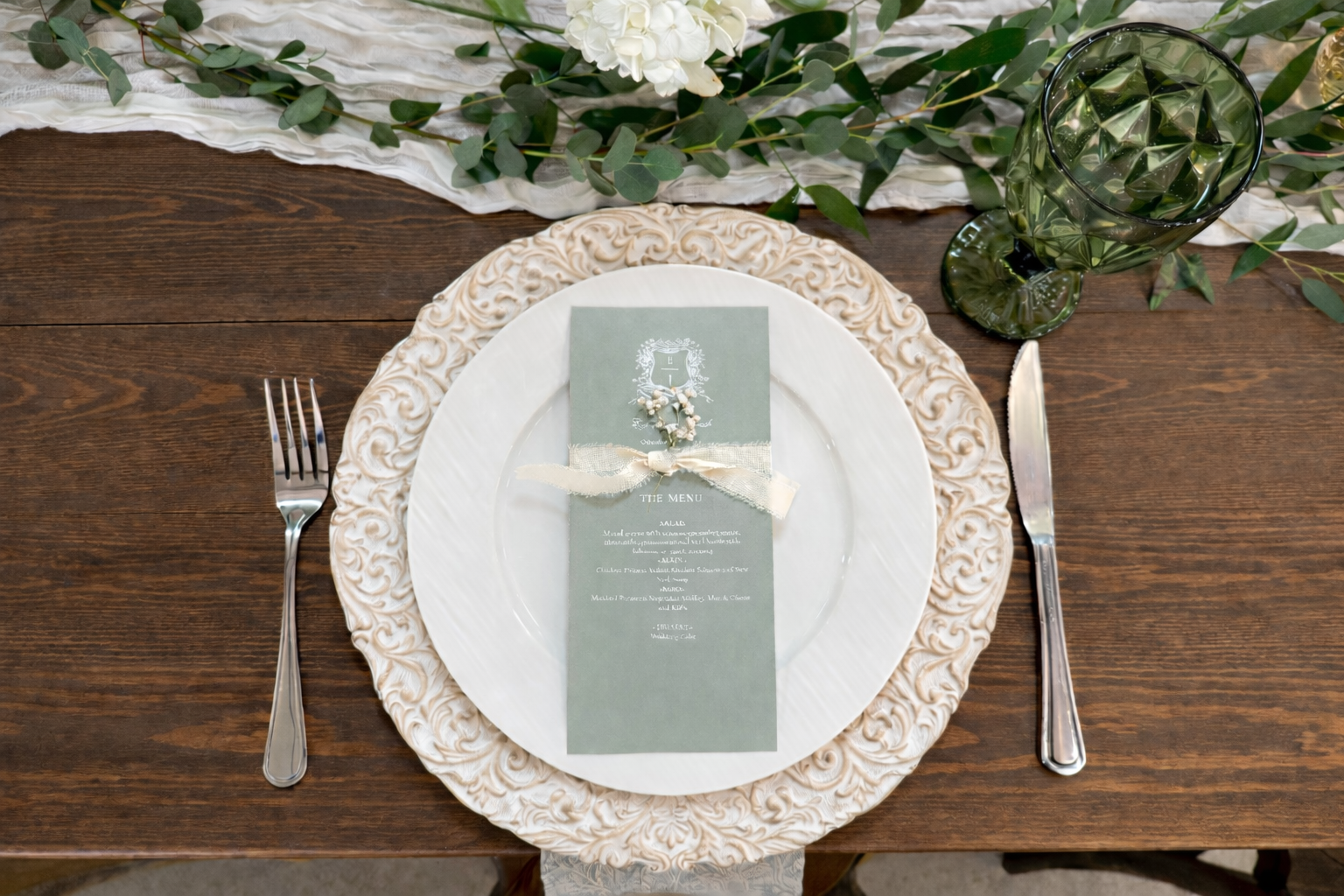 Wedding reception table setting featuring a decorative beige charger plate, white dinner plate, green menu card with a flower and ribbon embellishment, silver fork and knife, a green patterned drink glass, and greenery garland on a wooden table.