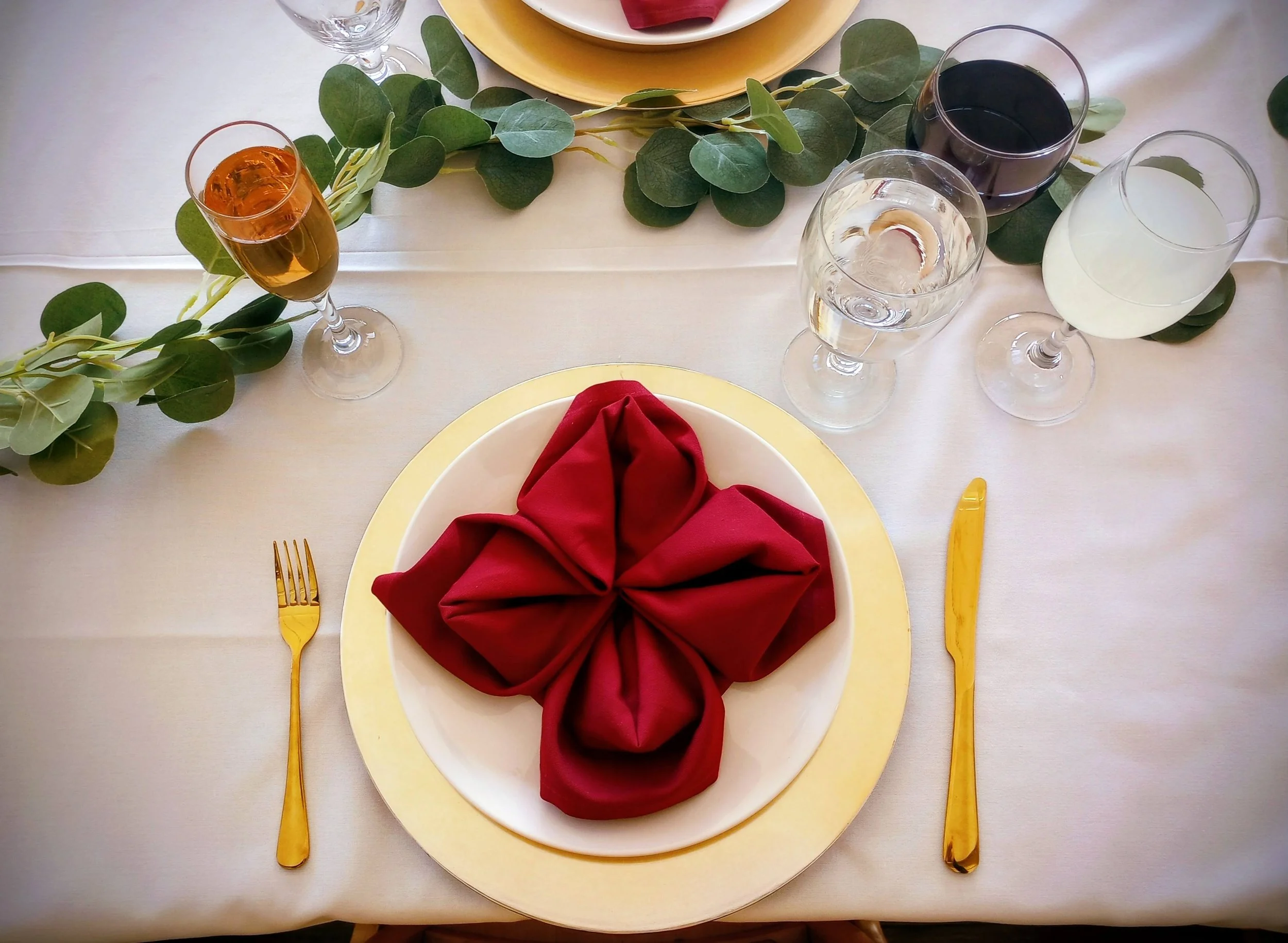 Table setting with a white tablecloth, a gold-rimmed plate with a red folded napkin in a flower shape, gold fork and knife, and four wine glasses filled with varied beverages, including red wine, white wine, water, and an amber-colored drink; green leafy garland runs across the table.