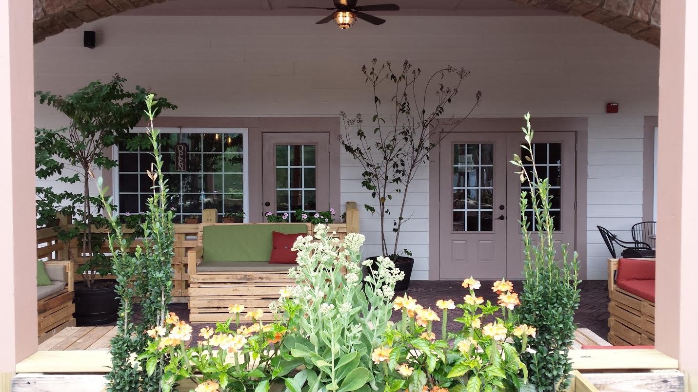 Outdoor patio with wooden furniture, green cushions, red throw pillows, and potted plants, including trees and flowering plants, against a white house with double doors.