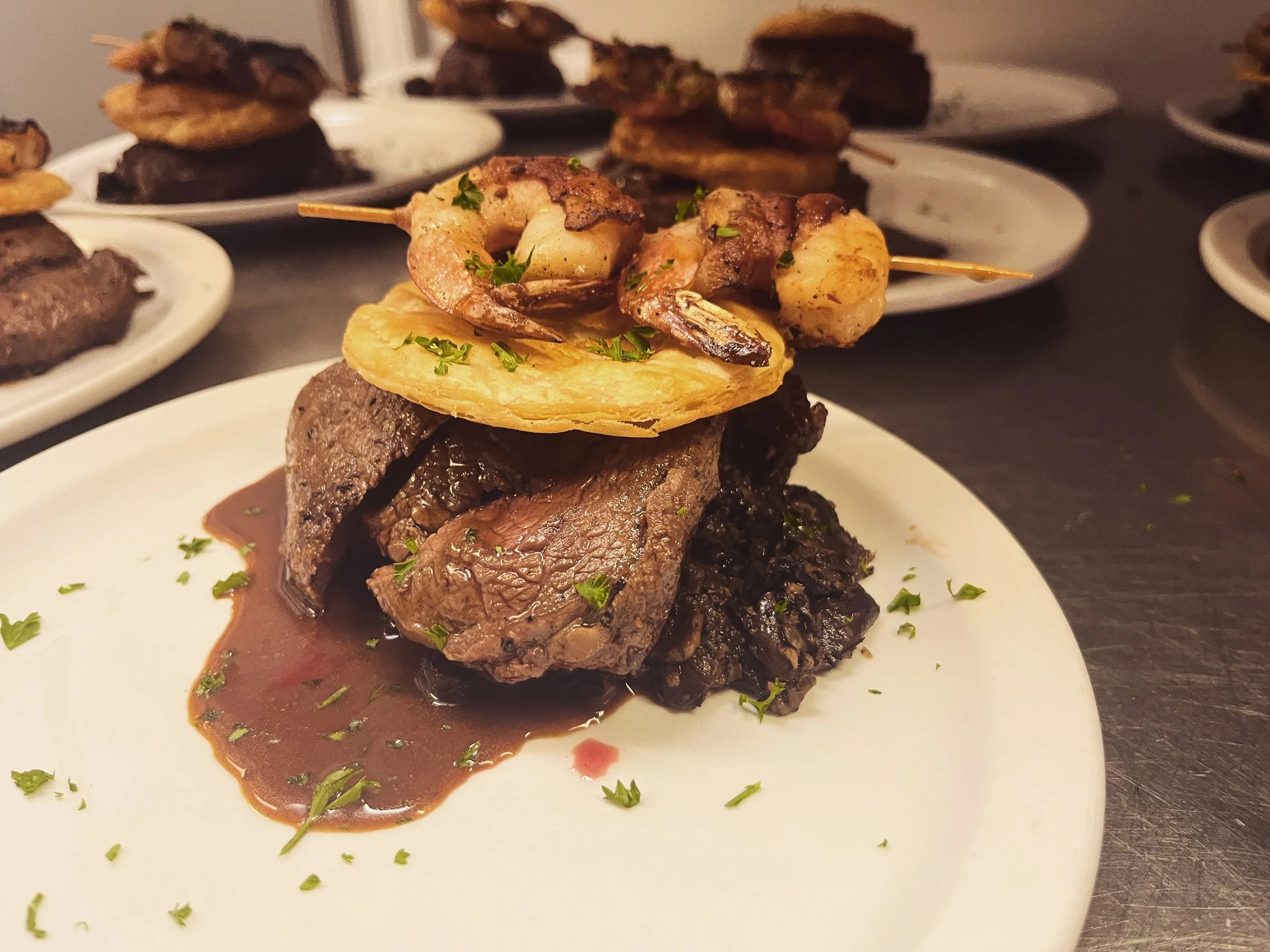 Elegant plated beef tenderloin with a mushroom and red wine reduction, topped with a shrimp skewer and a pastry round, garnished with chopped parsley.