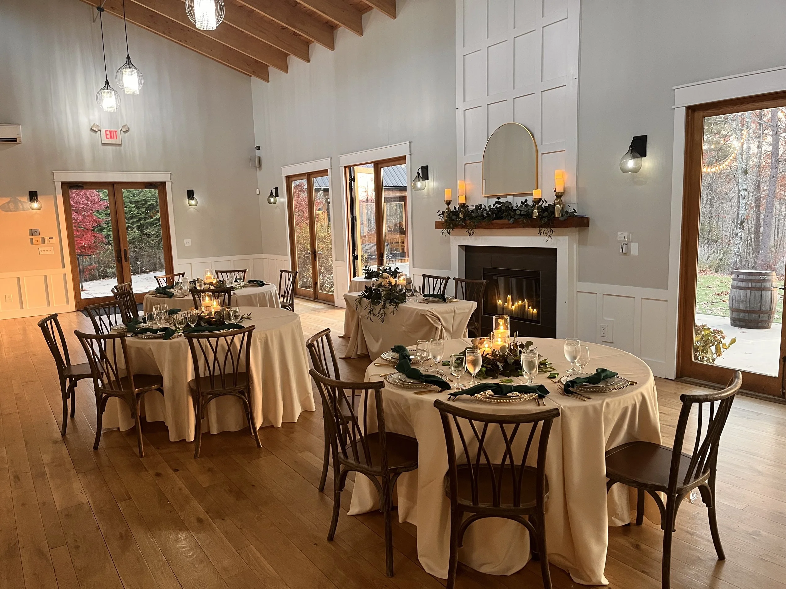 Elegant indoor dining room set for a special occasion with round tables covered in cream tablecloths, dark green napkins, glassware, and candles. The room features wooden floors, a white paneled wall, a fireplace decorated with greenery and candles, and large windows and glass doors looking outside at trees with autumn foliage.