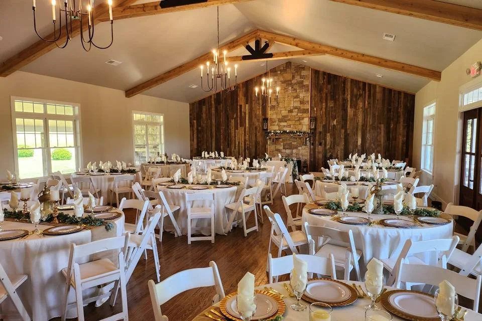 Wedding reception hall with round tables covered in white tablecloths, decorated with gold chargers and napkins, set for guests. The room features a high ceiling with exposed wooden beams, large windows providing natural light, a stone fireplace on the back wall, and chandeliers hanging from the ceiling.