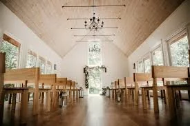 Interior of a bright dining area with wooden tables and benches, high ceiling with a chandelier, large windows, and natural light.