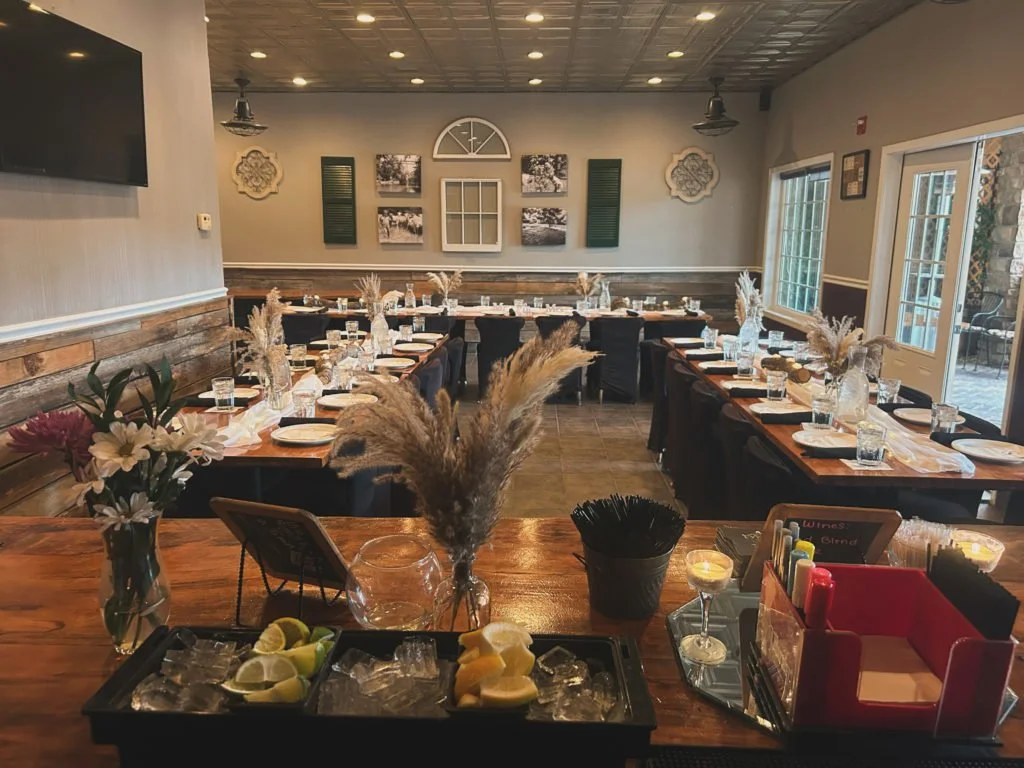 A restaurant dining room set up for a meal with tables arranged in a U-shape, decorated with vases of dried grasses, water glasses, and place settings. The space has wooden accents, framed wall art, and large windows.