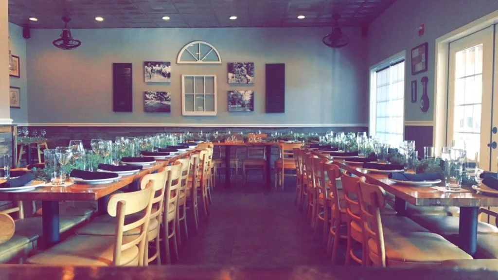 A dining room set up for a large gathering with long wooden tables, plates, glasses, and napkins, with chairs arranged along both sides. There is decorative greenery on the tables, and framed pictures and wall art on the walls. Sunlight enters throug
