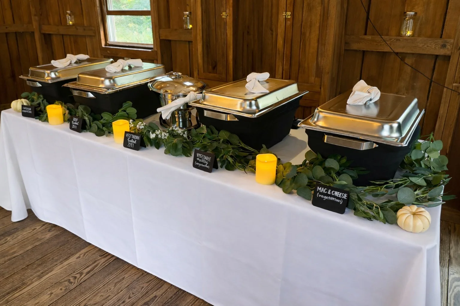 Buffet table with four chafing dishes, wrapped plates, and small chalkboard signs labeling dishes such as vegetable, macaroni and cheese, and chicken. Greenery and small candles decorate the table, which is covered with a white tablecloth.
