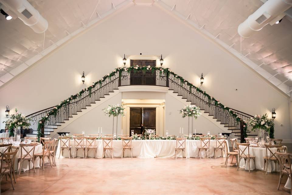 Elegant wedding reception hall with long head table, floral centerpieces, and decorated staircase.
