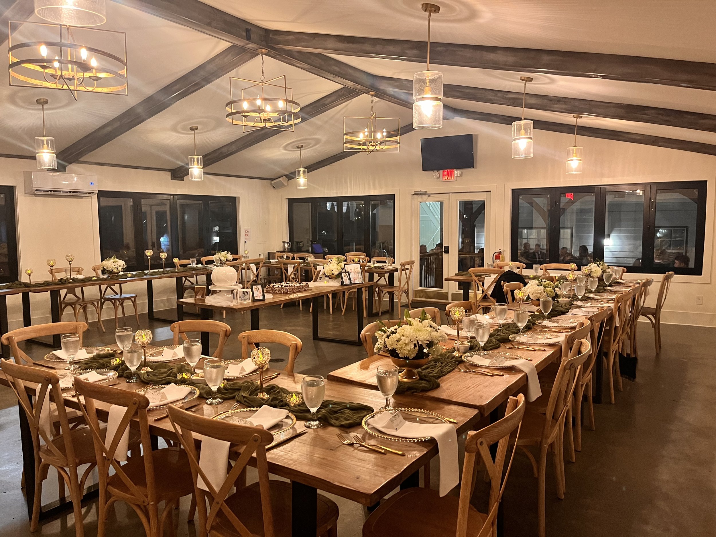 Decorated dining room with long wooden tables set with white napkins, glasses, and floral centerpieces, arranged for a formal event.