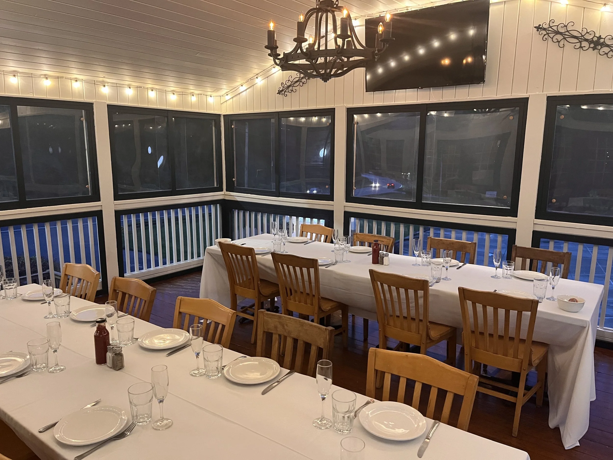 Empty dining area with long tables covered with white tablecloth, set with plates, utensils, and glasses, surrounded by wooden chairs, enclosed by large windows, decorated with string lights and a chandelier, in a cozy, rustic style.